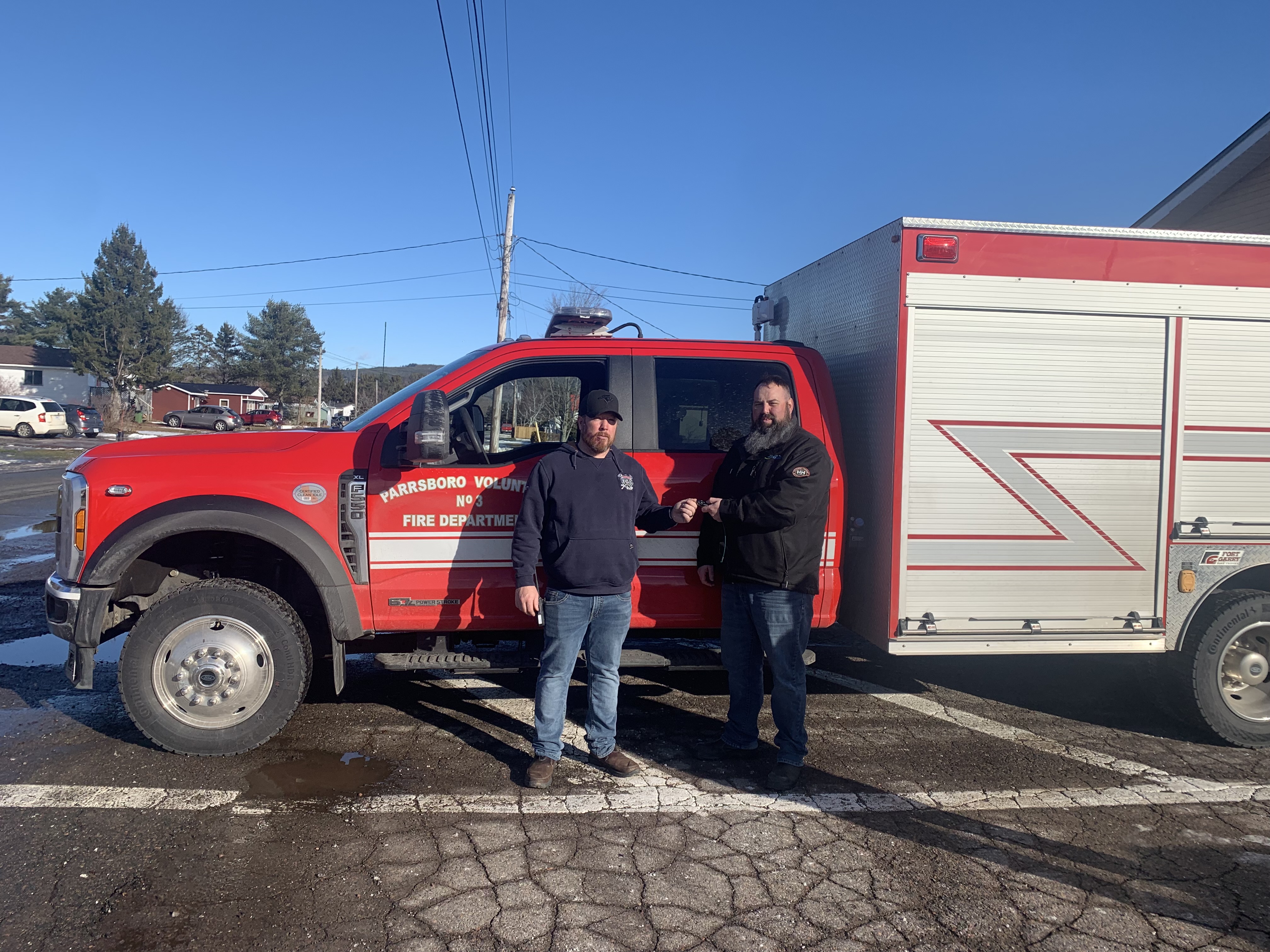 Two men stand in front of a red fire-rescue vehicle on a sunny day.